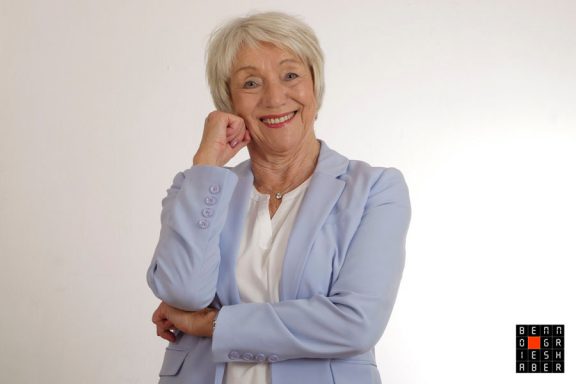 Lächelnde ältere Frau in blauer Jacke, mit der Hand am Kinn, vor hellem Hintergrund.