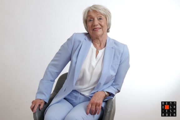 Ältere Frau mit hellen Haaren, lächelnd, in einer blauen Outfit, sitzt auf einem Stuhl.