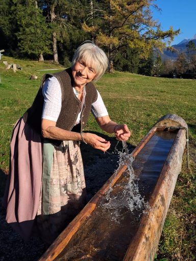 Ältere Frau füllt Wasser aus einem Holzbrunnen in einer ländlichen Umgebung.